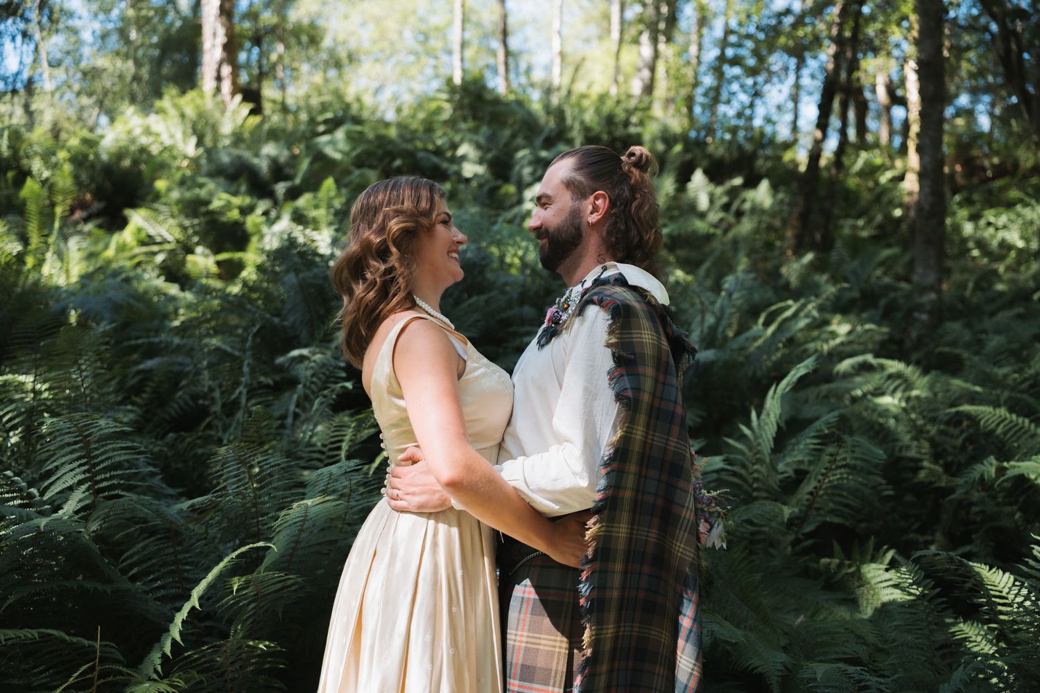Wedding couple poses on beach shoreline with misty lake view, groom wearing traditional Scottish kilt.