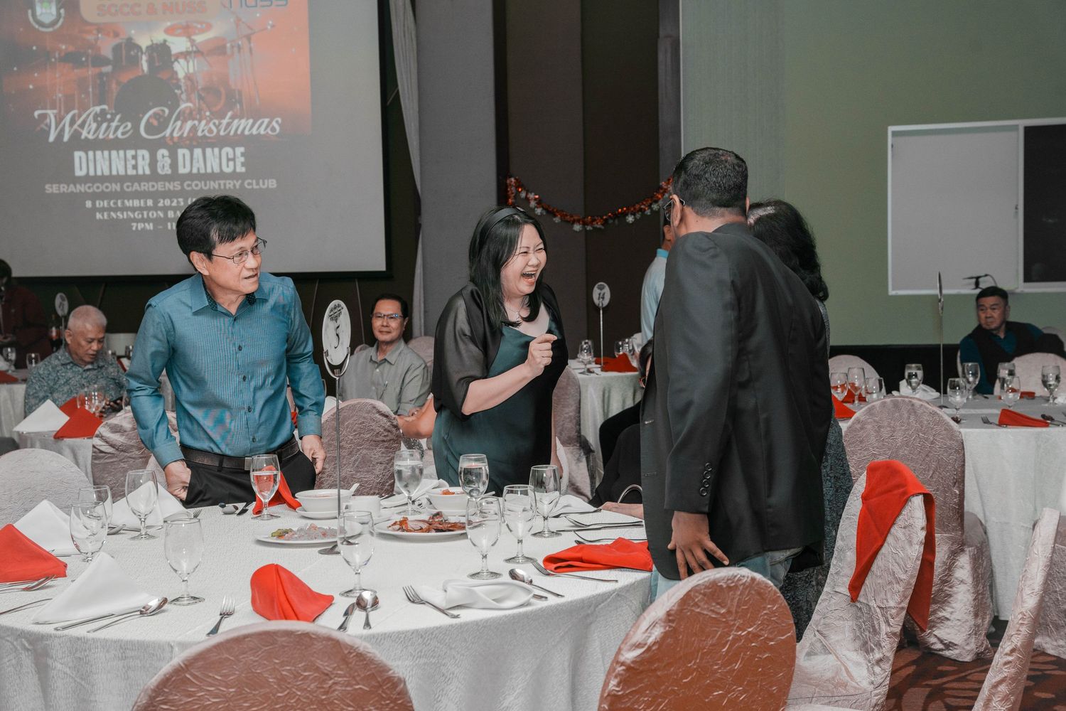 Guests mingling at elegant dinner tables decorated with white linens and red napkins at a formal event.