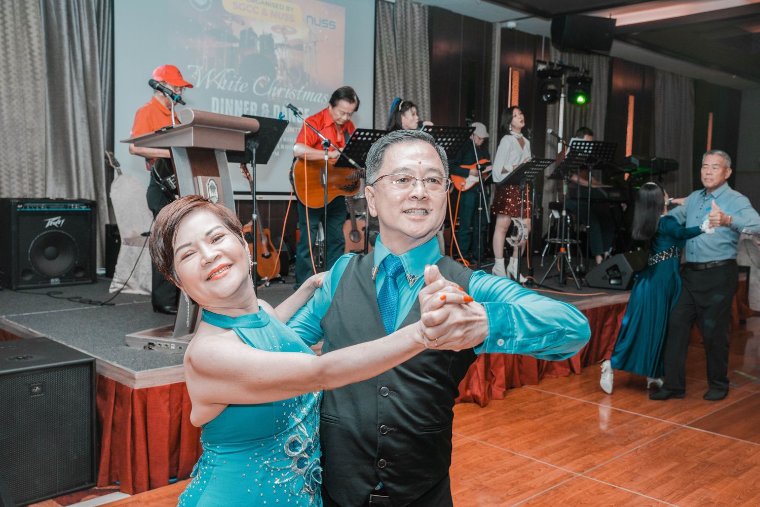 Couple dancing in turquoise attire with live band performing on stage in background.