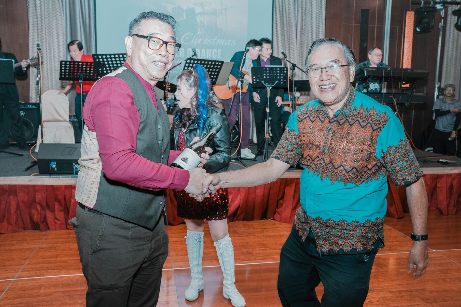 Two guests sharing a friendly handshake at a formal event with red stage decorations in the background.