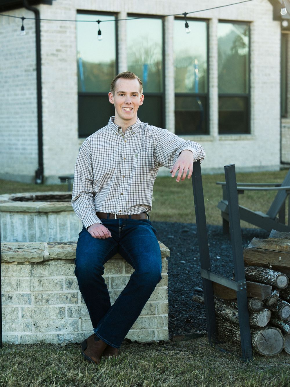 Person in checkered shirt sitting on brick ledge near outdoor firepit.