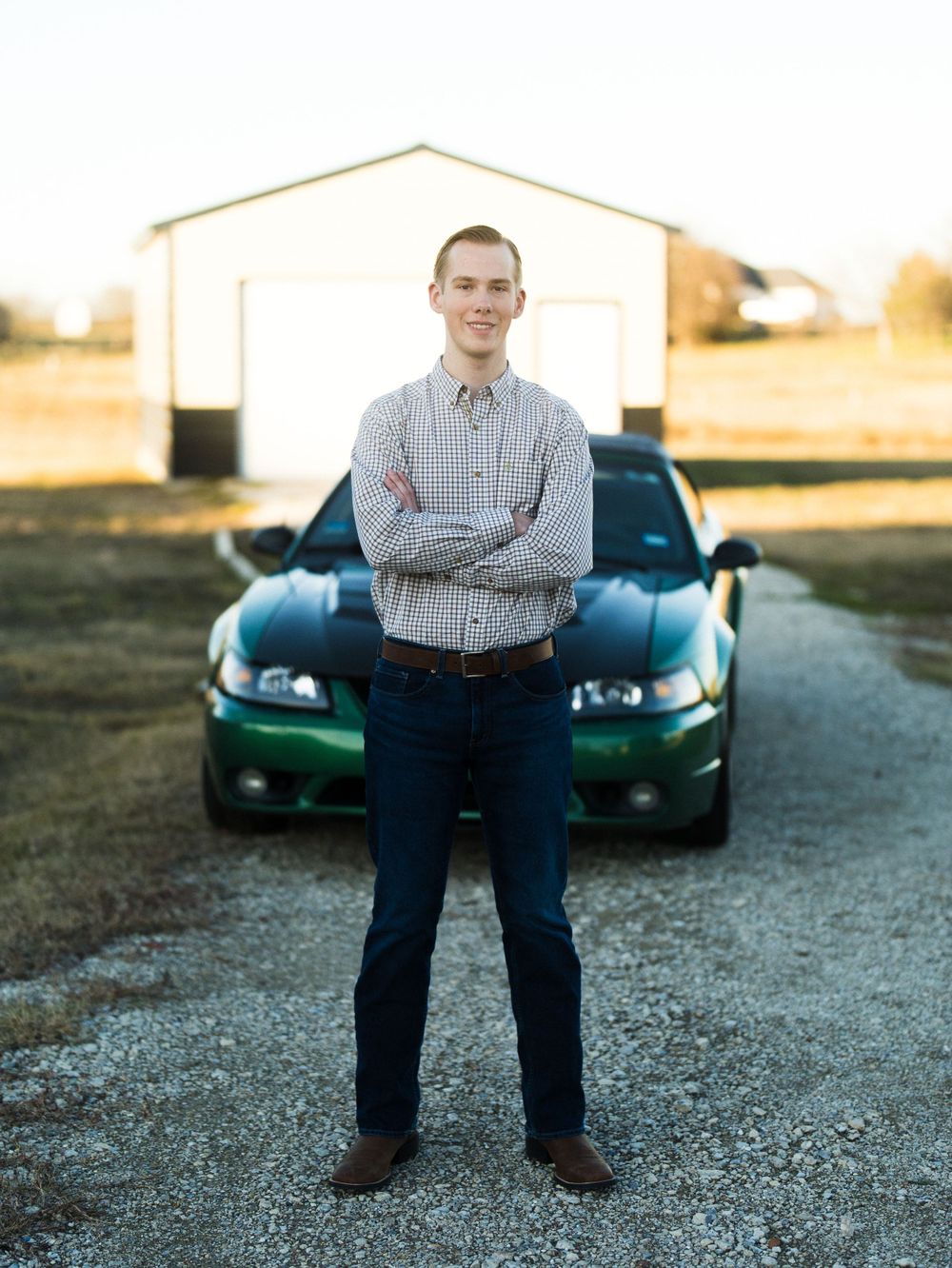 Person in plaid shirt standing with crossed arms in front of green car and white building.