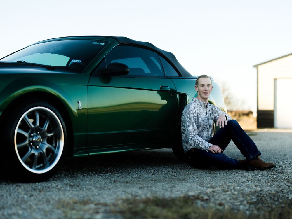 Dark green sports car parked at sunset with figure sitting casually beside it on the ground.