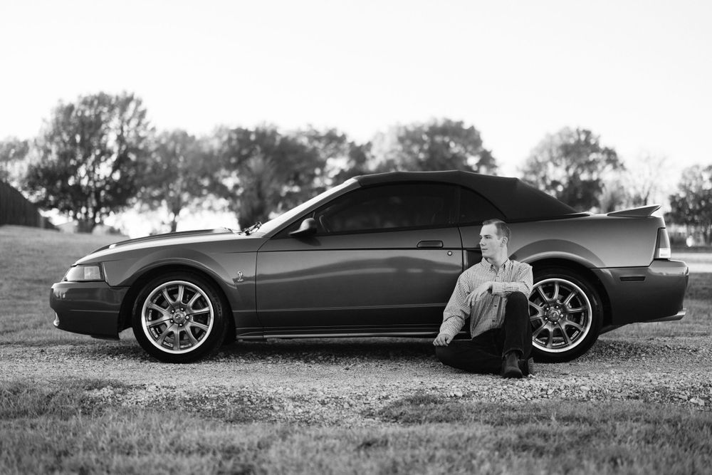Black and white photo of a sleek sports car with custom wheels against a rural backdrop.