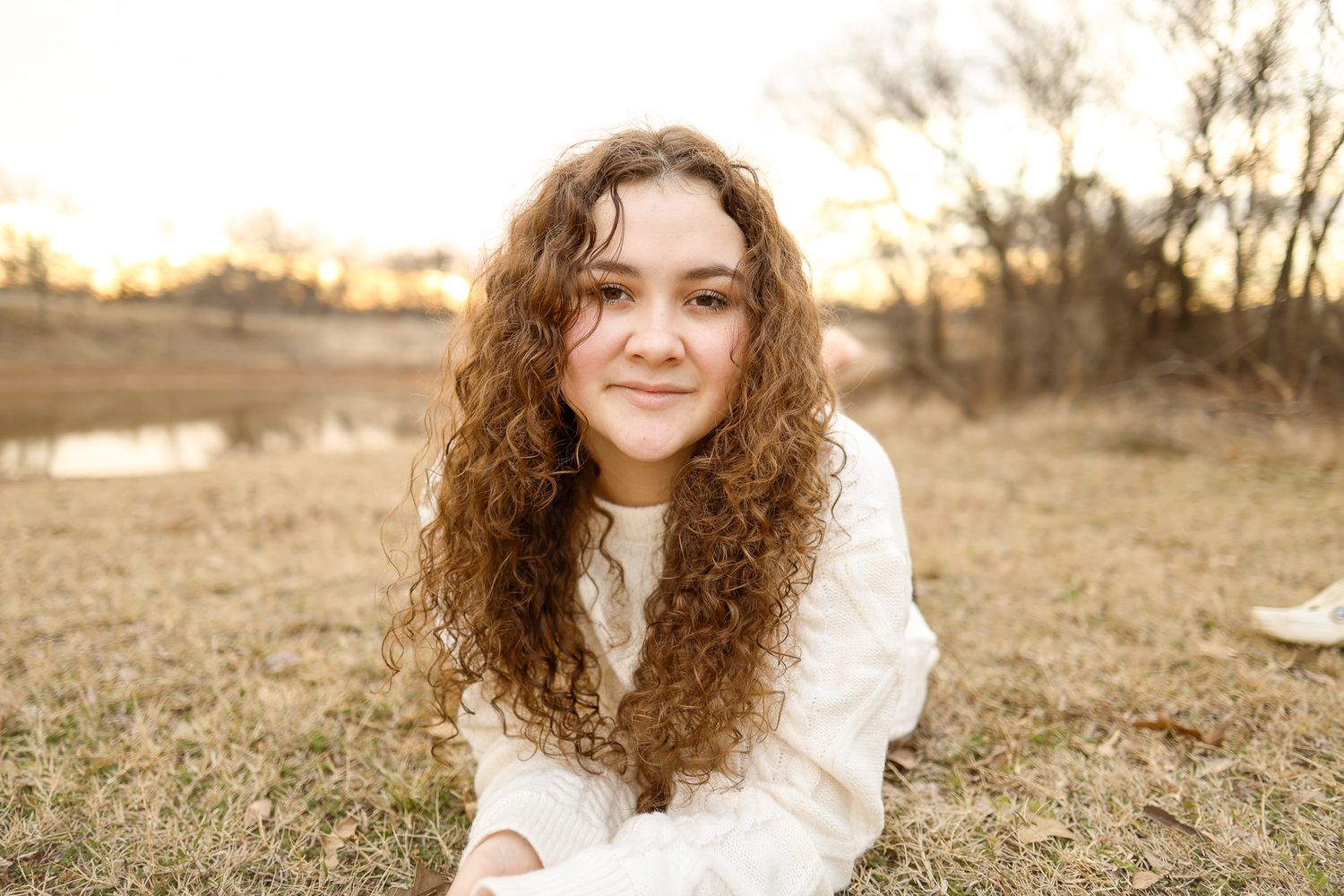 Outdoor portrait against golden sunset background featuring someone in white sweater with curly hair.
