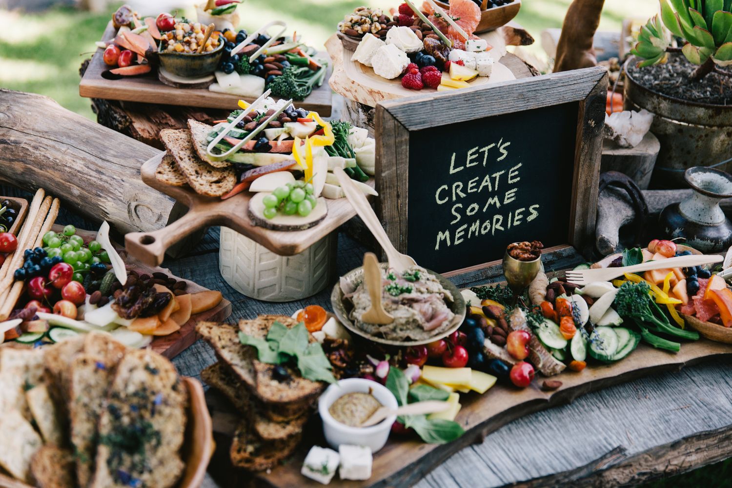 Rustic outdoor grazing table spread with charcuterie, fruits and sign saying Let's Create Memories on wooden boards.