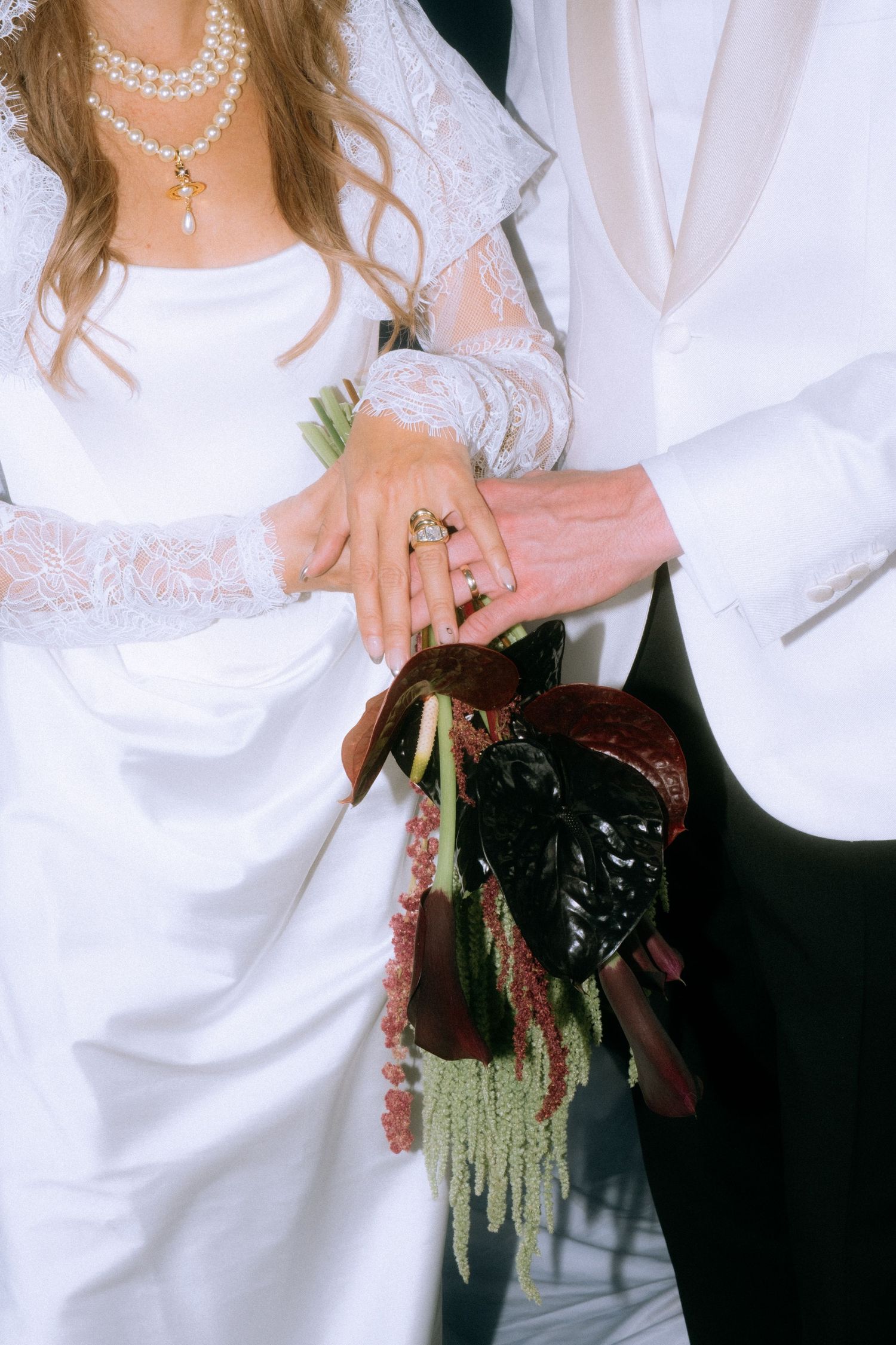 Wedding detail showing hands with rings, pearl necklace, and white lace sleeves holding a bouquet.