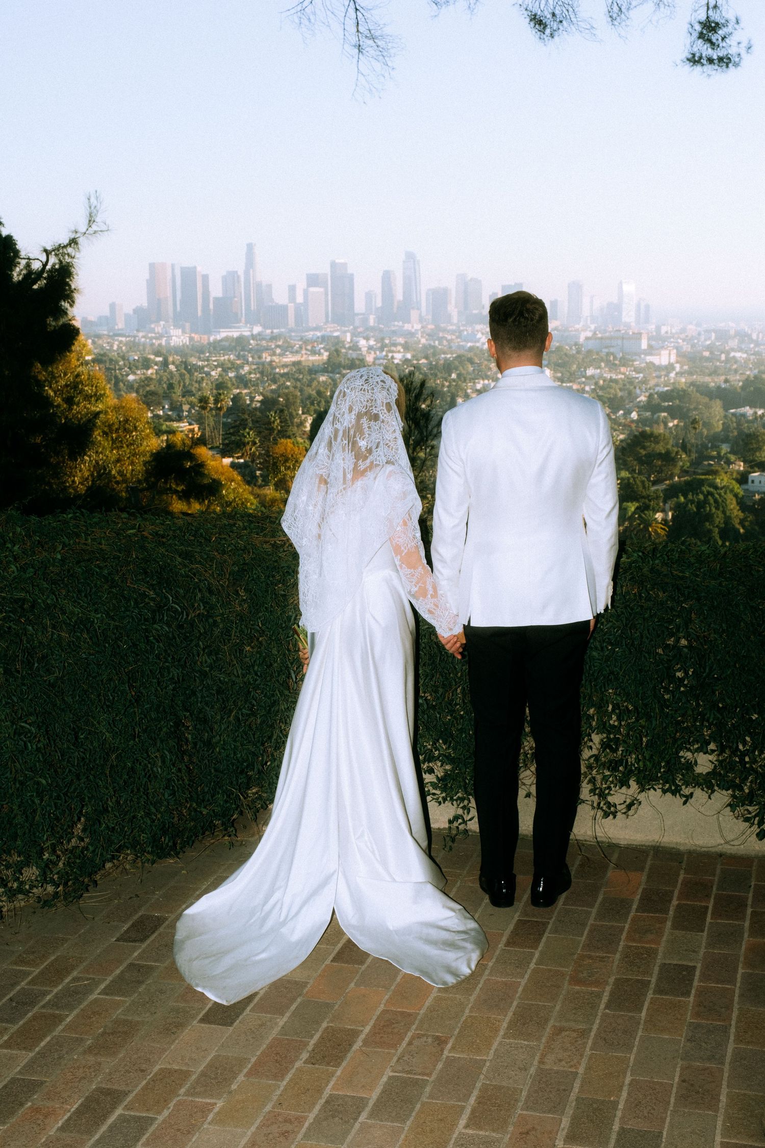 Couple in wedding attire viewing city skyline from hilltop brick pathway.