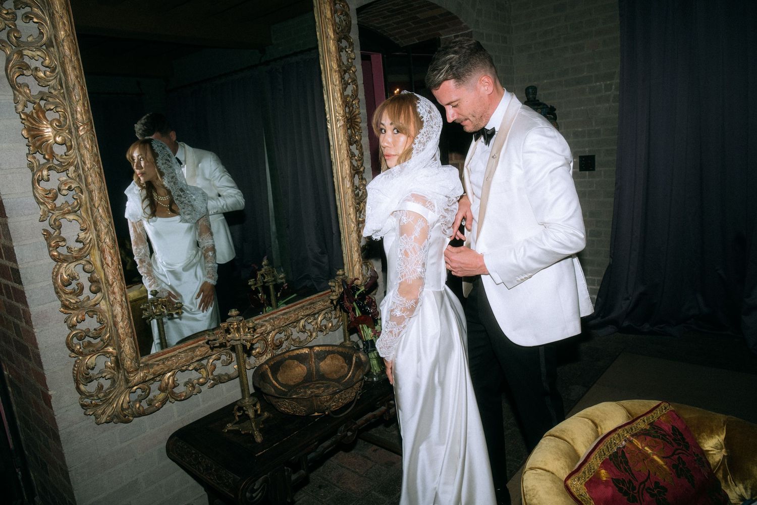Elegant couple in formal wedding attire posing near ornate vintage mirror.