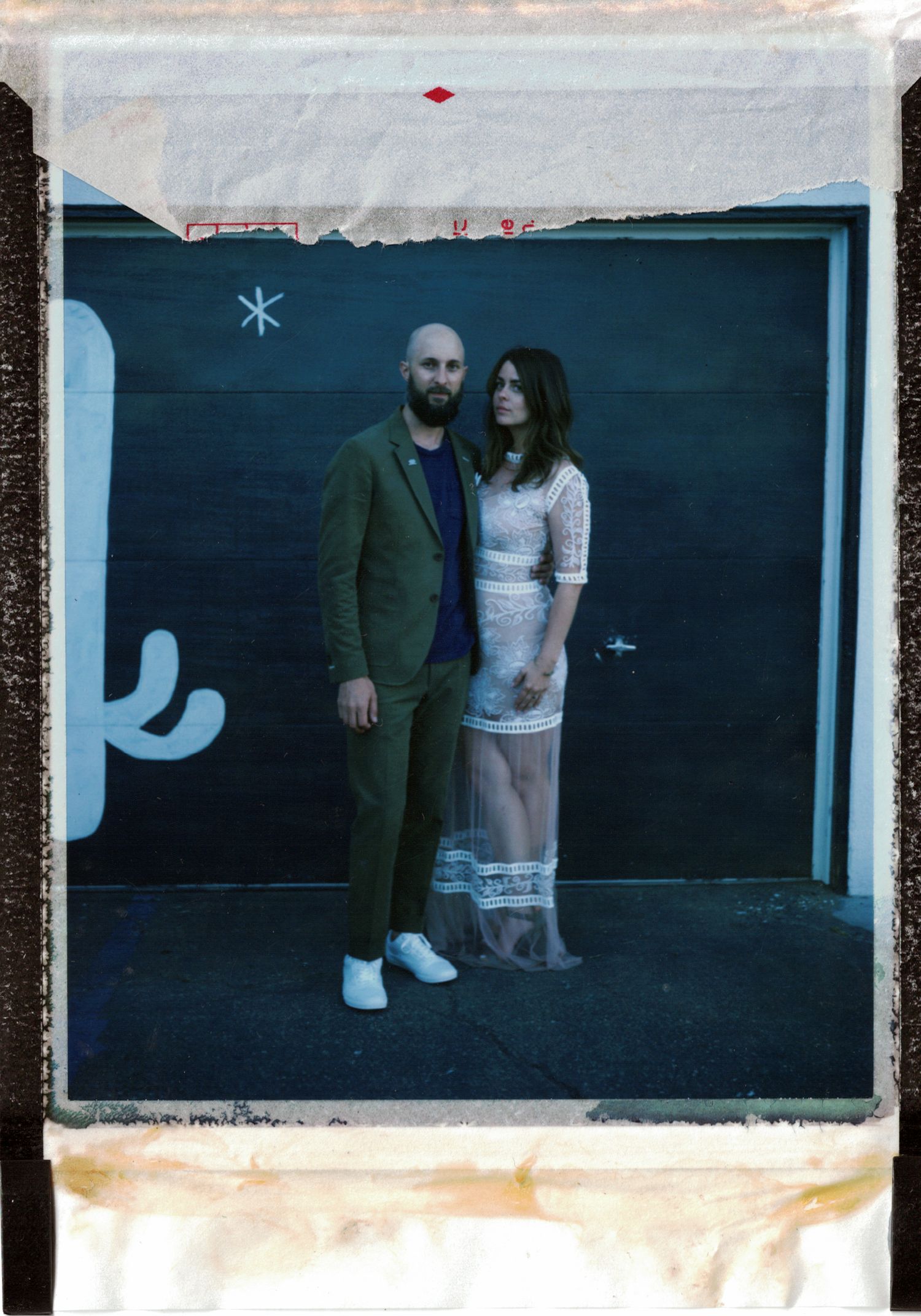 A couple poses together against a dark wall with the person on the right wearing a white lace dress and left in olive suit.