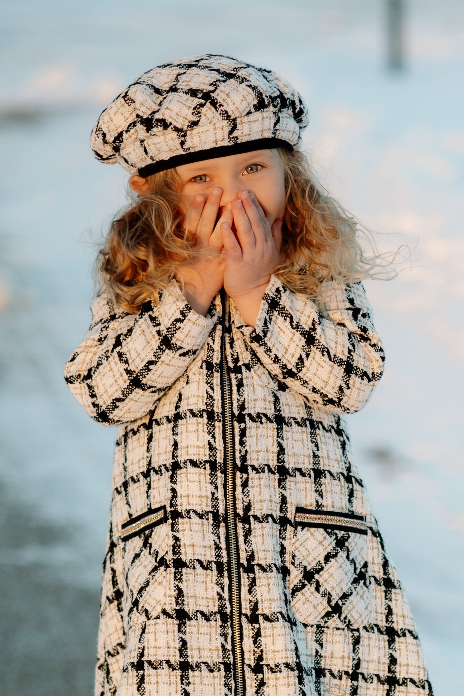 Child in black and white plaid coat and matching hat giggles while covering mouth.