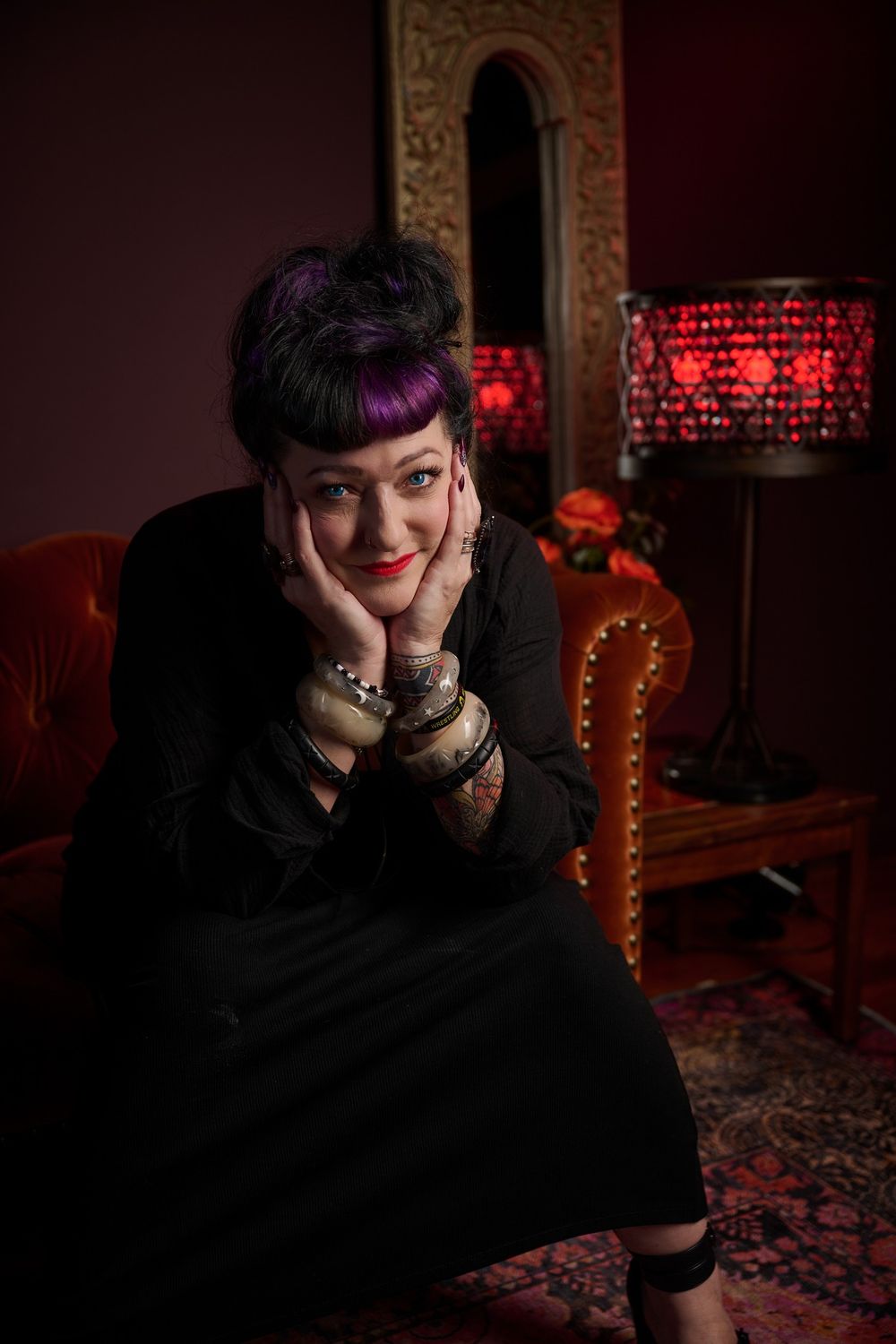 Someone with dark hair and bangs poses dramatically in moody red lighting against an ornate interior setting.