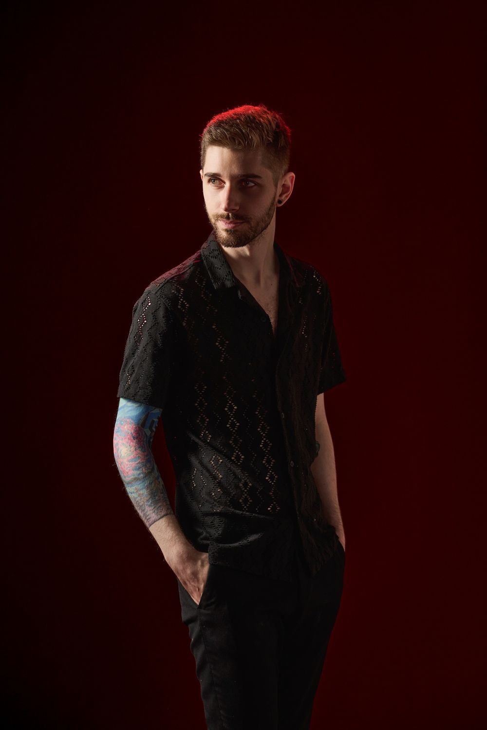 Dramatic studio portrait against dark red background featuring individual in black clothing with colorful arm tattoos.