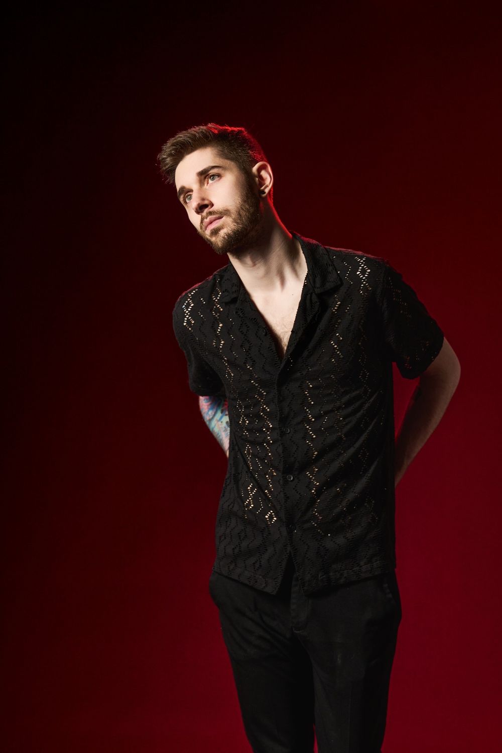 Fashion portrait of someone wearing a black lace button-up shirt against a dark red background.