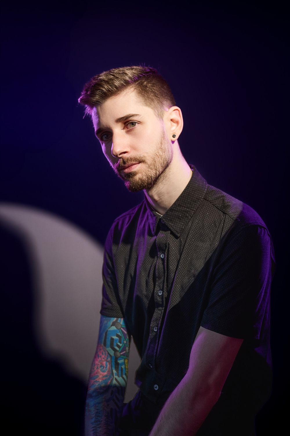 A performer in a black vest with tattoos on their arm poses against a dark background with purple lighting effects.