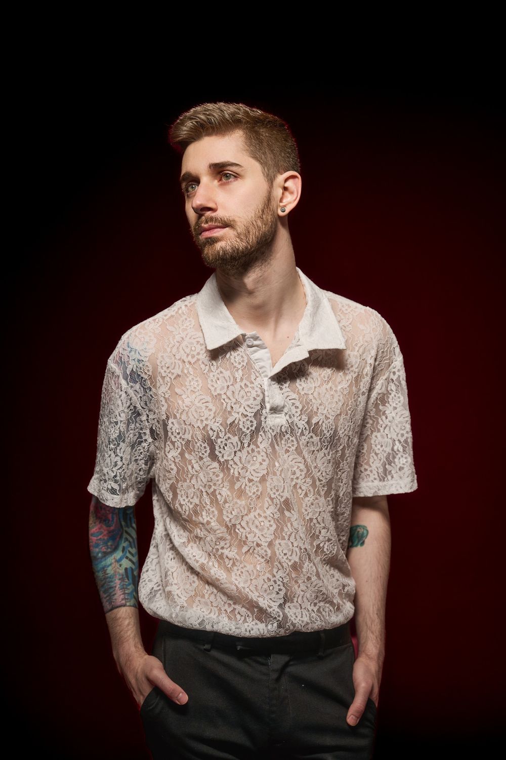 A person wearing a white lace polo shirt and dark pants poses against a dark red background.
