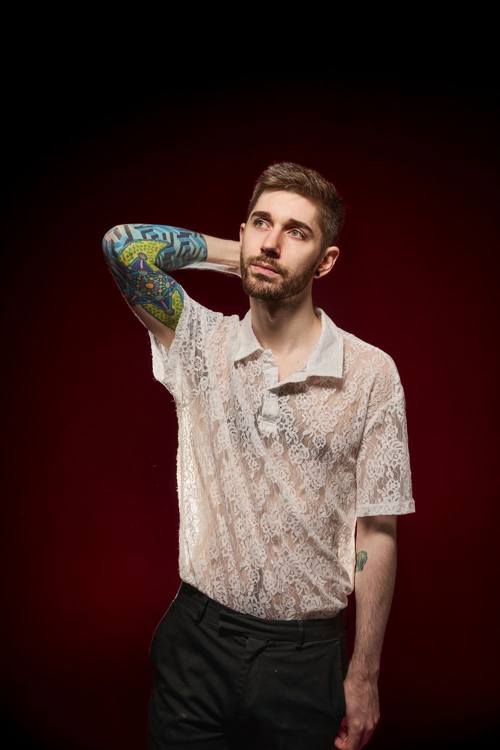 Person wearing a white lace polo shirt and dark pants poses thoughtfully against a dark red background.