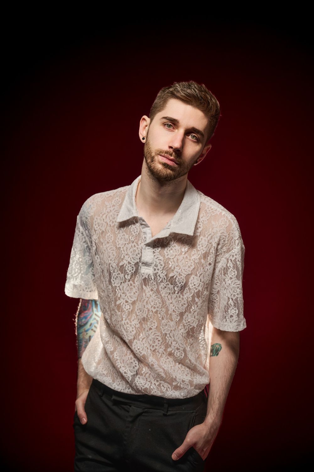Portrait against dark red background showing figure in white lace polo shirt with visible tattoos and one hand in pocket.