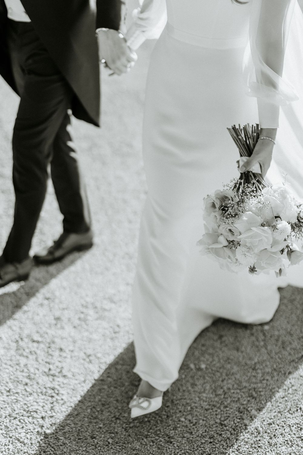 Black and white detail shot of wedding dress and bouquet while couple holds hands.