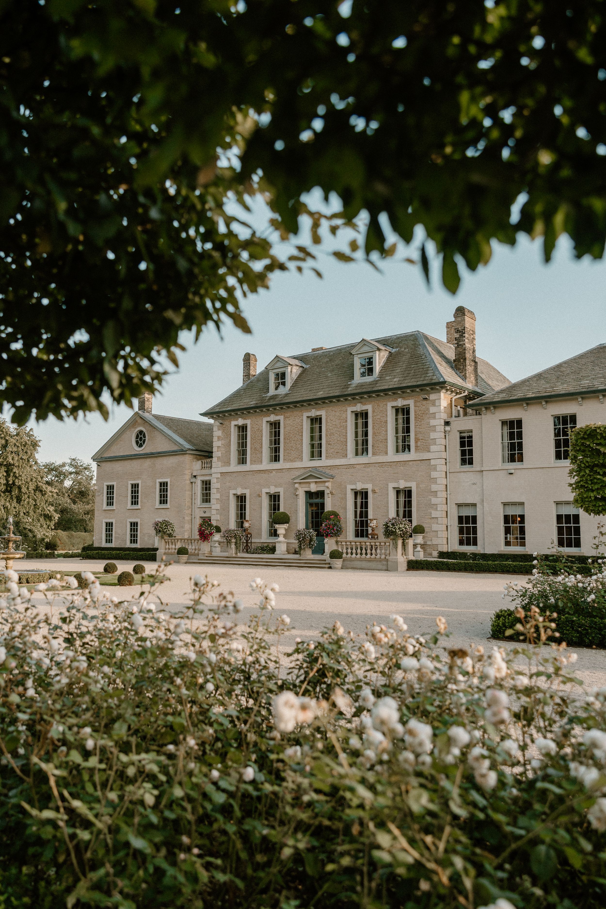 Elegant Georgian-style mansion with white flowers in the garden and tree framing the entrance.