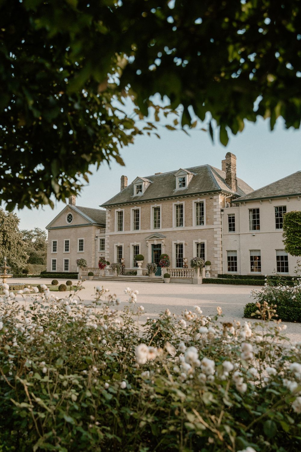 Elegant Georgian-style mansion with white flowers in the garden and tree framing the entrance.