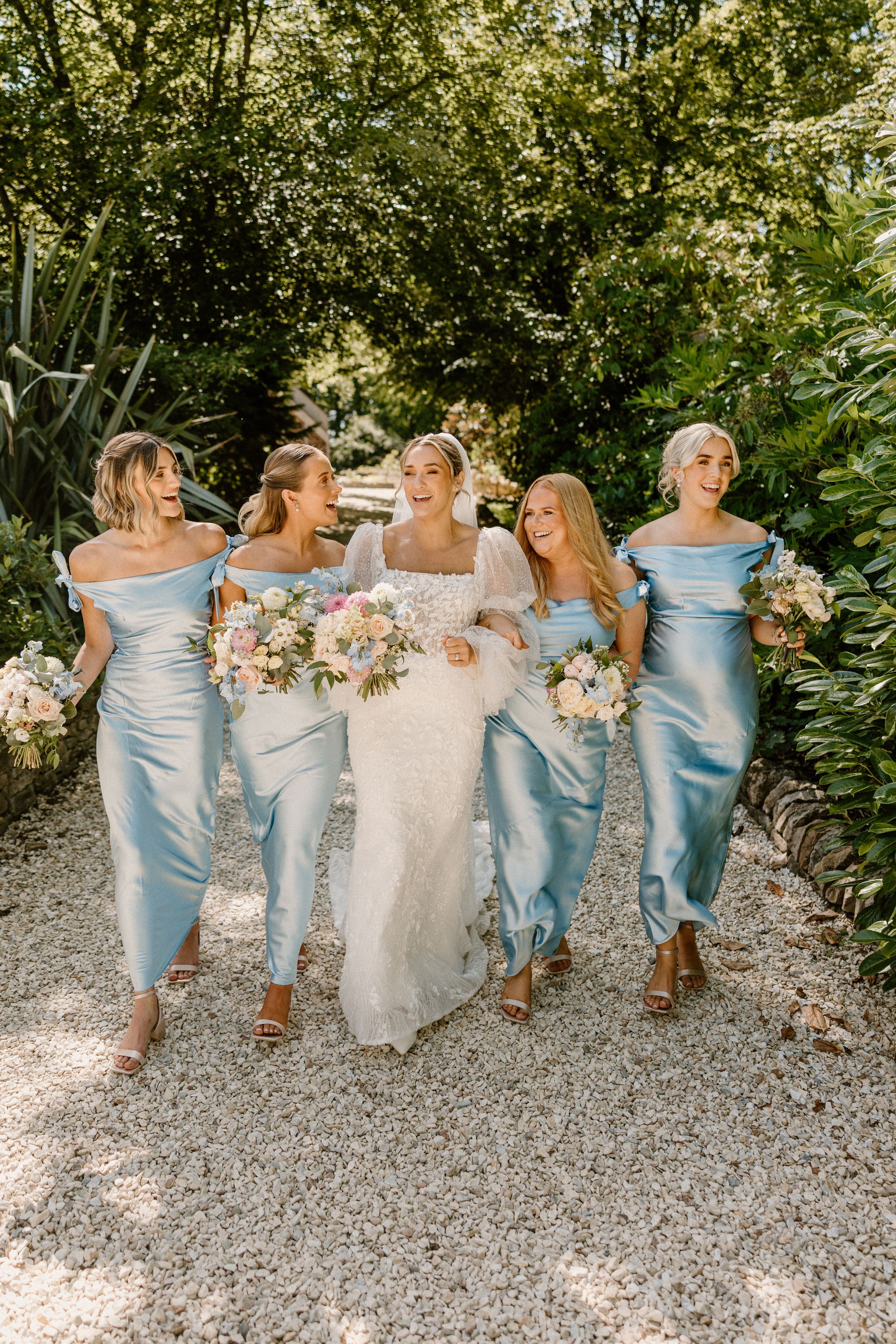 Bridesmaids in pale blue dresses sit side by side with bride in white dress on gravel path at outdoor wedding.
