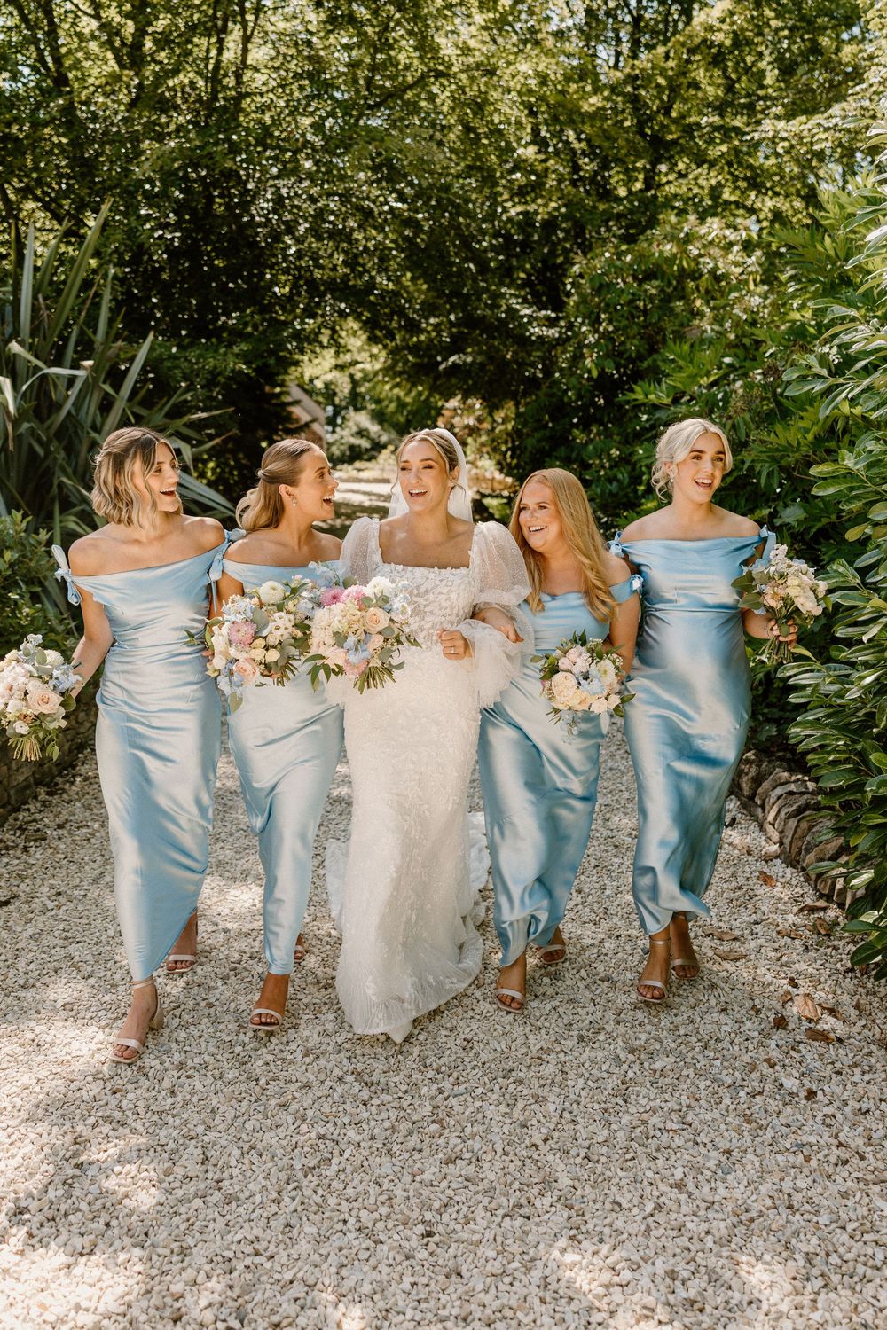 Bridesmaids in pale blue dresses sit side by side with bride in white dress on gravel path at outdoor wedding.