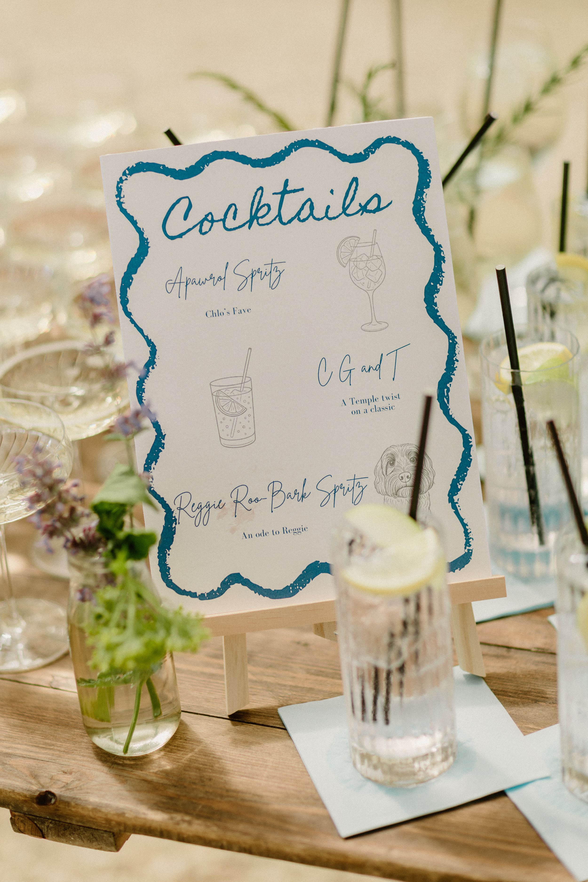 A rustic cocktail menu sign with blue border stands on a wooden table with glassware and greenery decorations.
