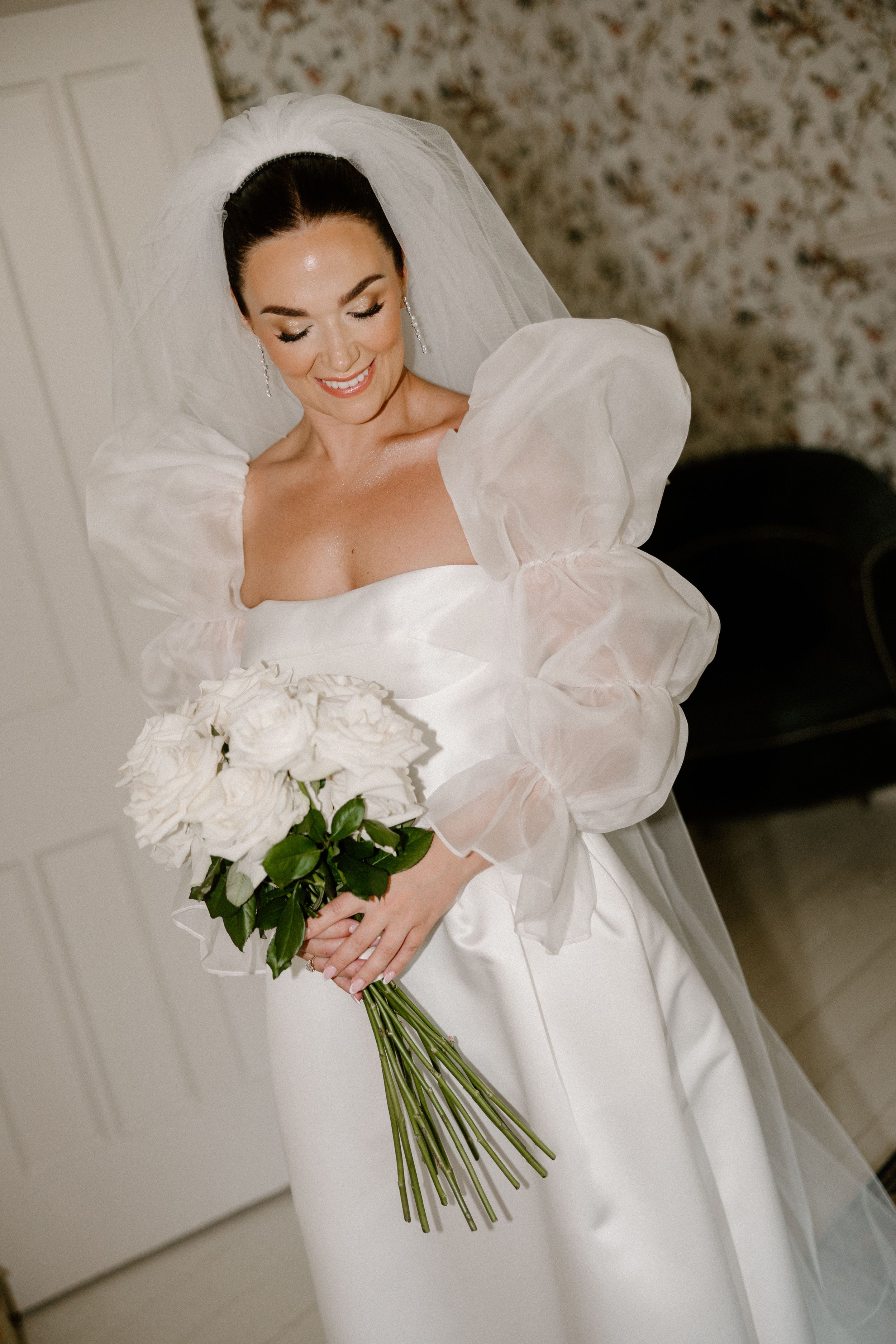Elegant bride in dramatic puff sleeve wedding dress holds white peony bouquet while smiling down.