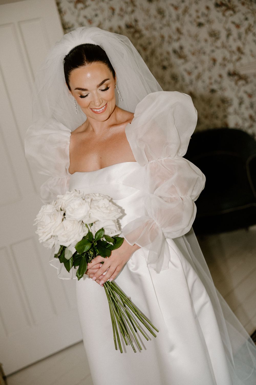 Elegant bride in dramatic puff sleeve wedding dress holds white peony bouquet while smiling down.