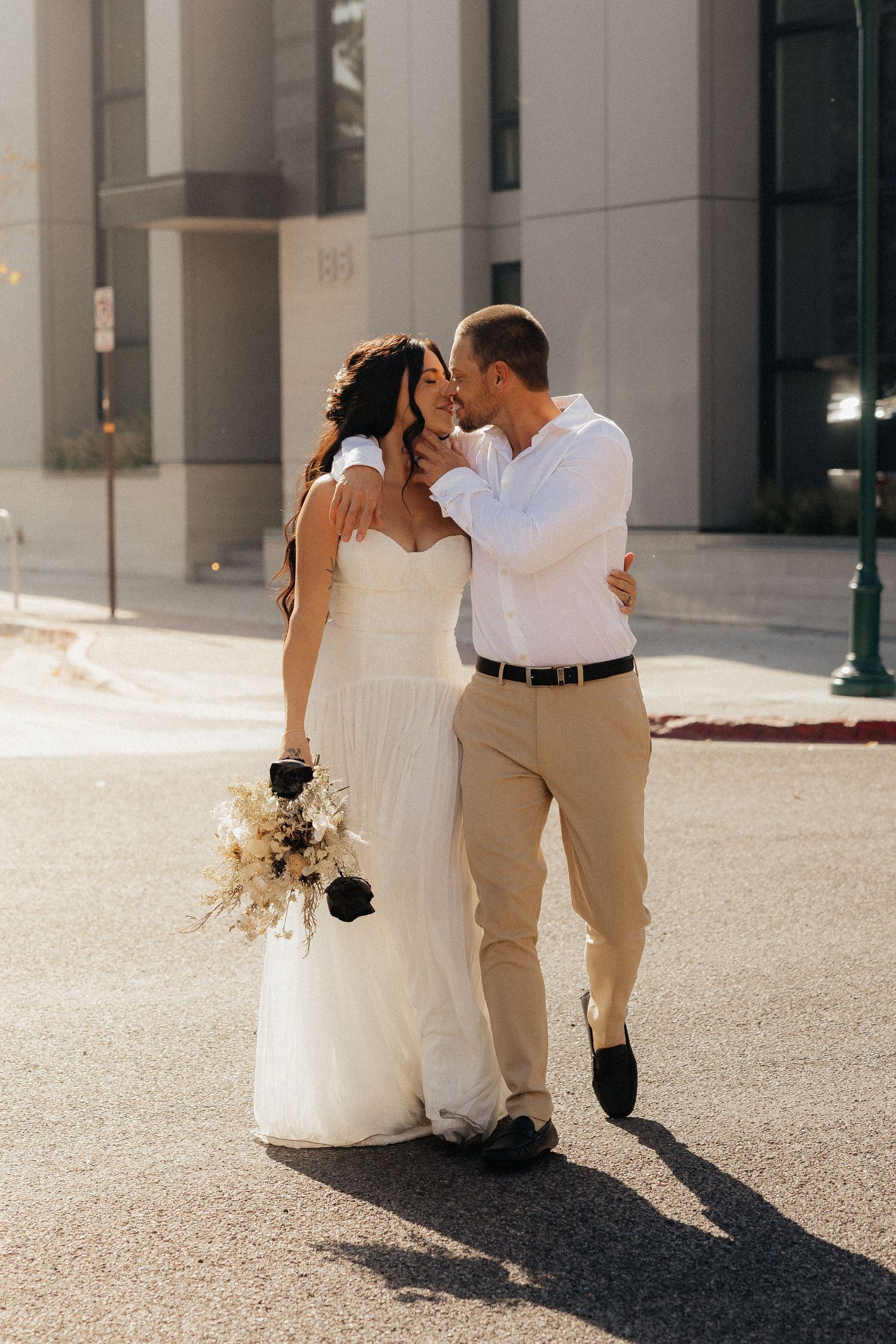 Sun-drenched wedding photo with dried flower bouquet on city street.