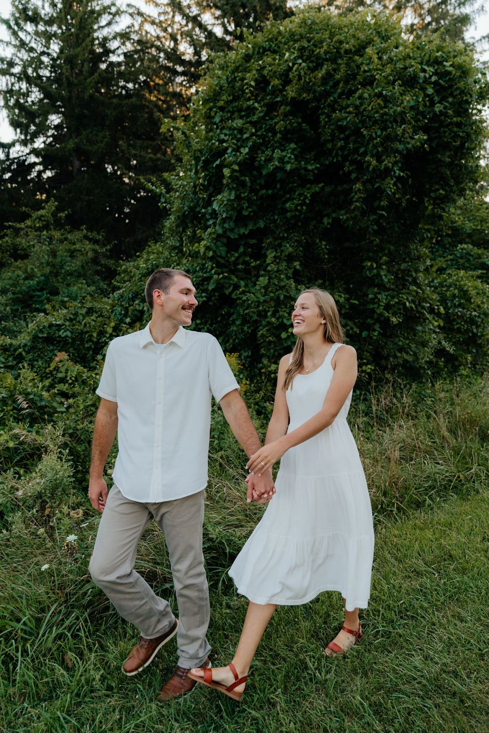 A couple in white summer outfits walks together on grass with lush greenery in the background.