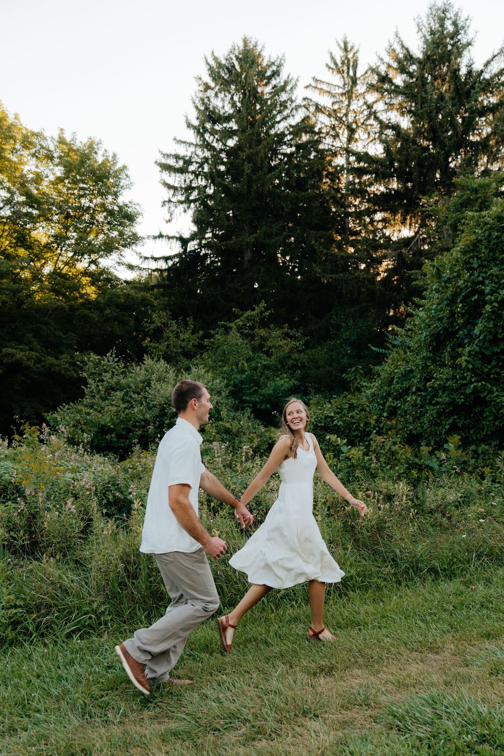Playful couple dances together on a grassy path surrounded by evergreen trees.