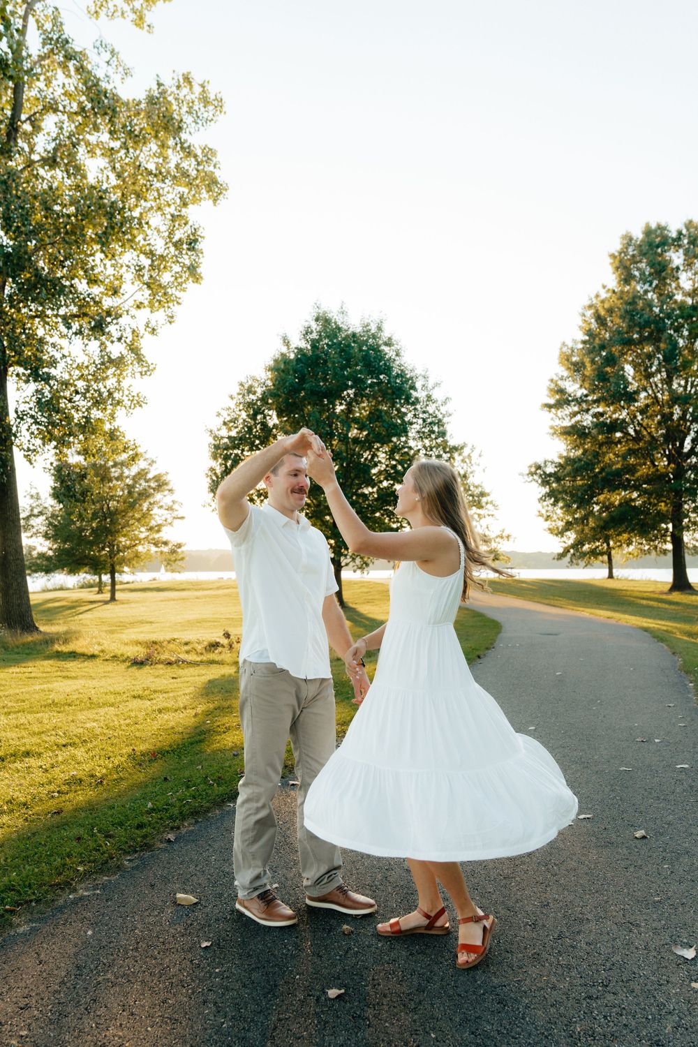 Couple dancing and twirling in white outfits on a tree-lined path.