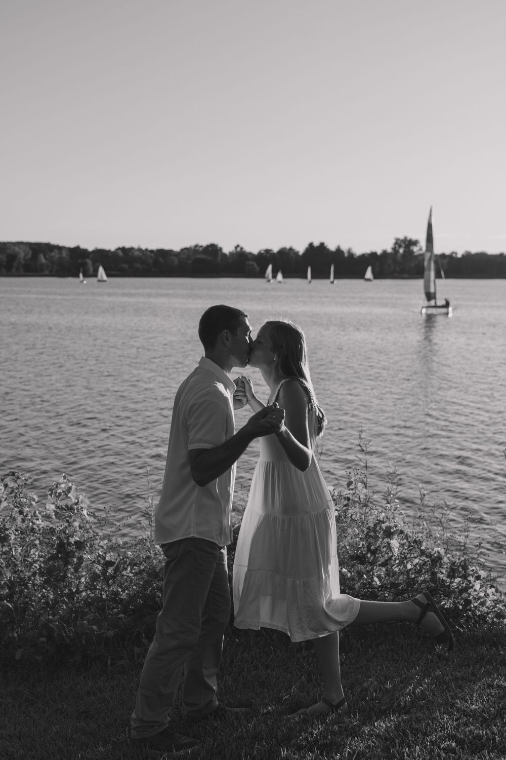Silhouetted couple by lake with sailboats in background at sunset.