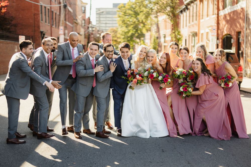Wedding party in grey suits and mauve dresses poses on a brick-lined city street.