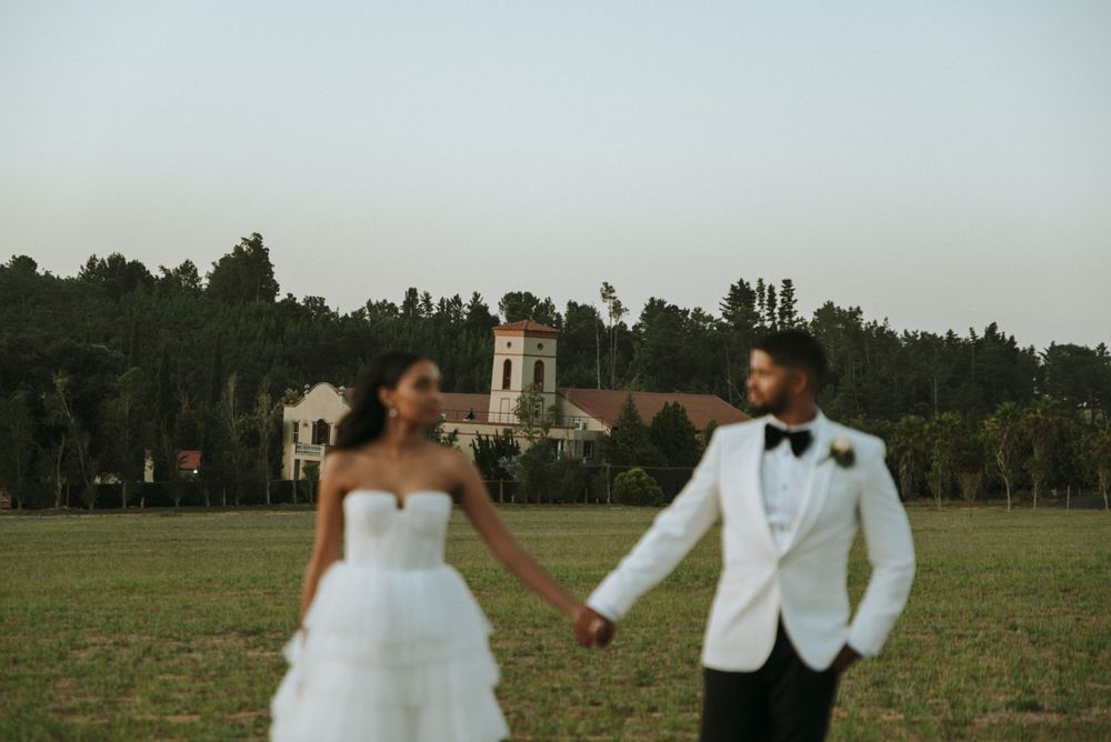 Newlyweds holding hands walking through estate grounds with buildings in background.