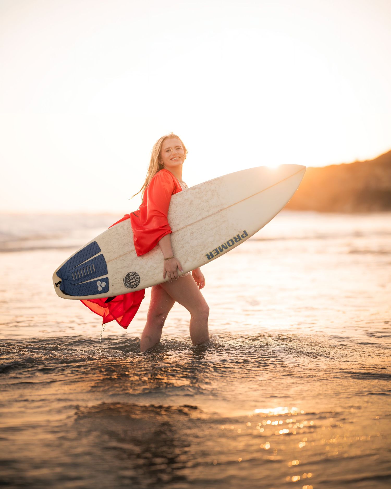 a female graduate in an orange gown holding a surfboard in the crystal cove sunset