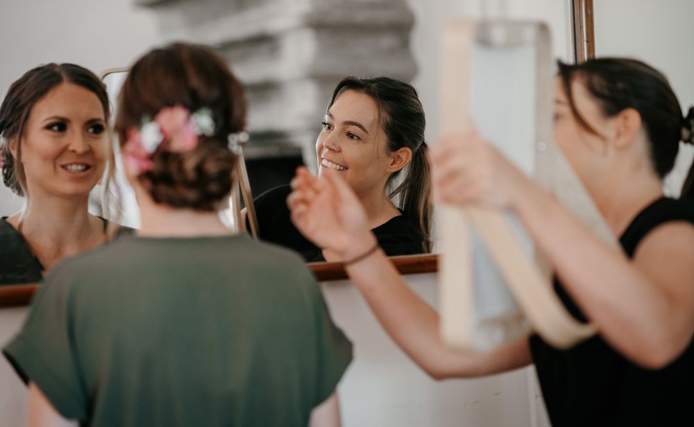 Two women smiling at each other in a mirror reflection during wedding preparation.