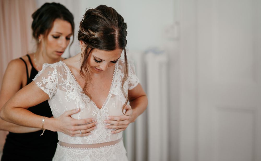 Bridesmaid helping bride fasten her lace wedding dress from behind.