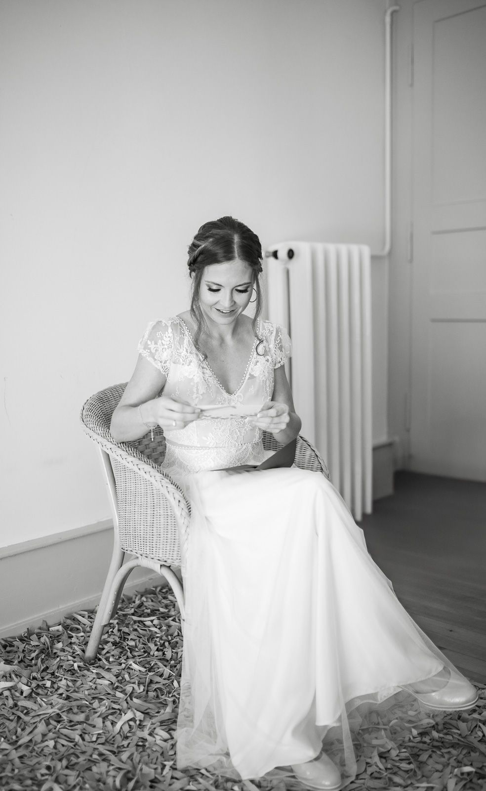 Bride in lace wedding gown sitting in wicker chair reading a letter, black and white photo.