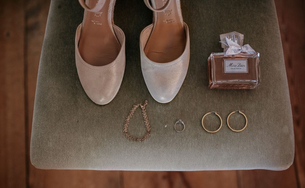 Flat lay of bridal accessories including nude heels, gold jewelry, and Miss Dior perfume on velvet surface.