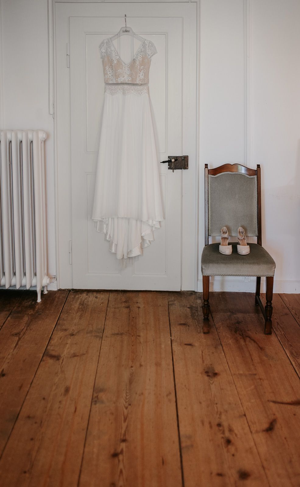 Elegant lace wedding dress hanging on white door beside vintage chair with bridal heels inside.