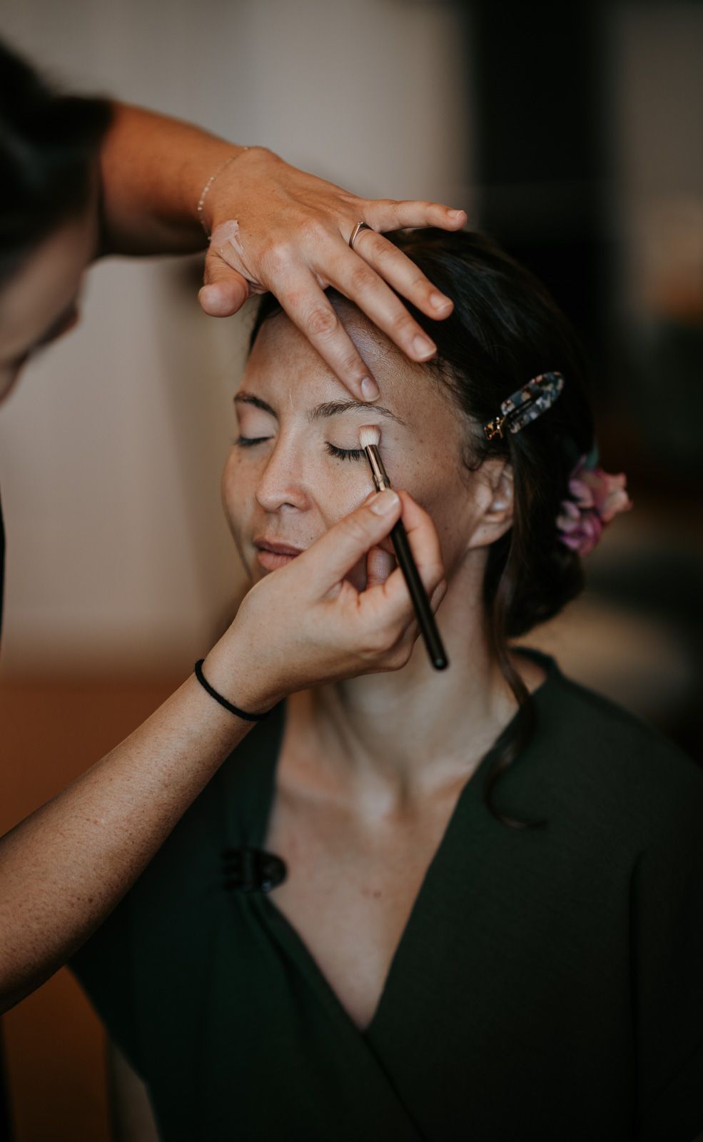 Makeup artist applying eyeshadow to a woman in a green jumpsuit with hair clip.