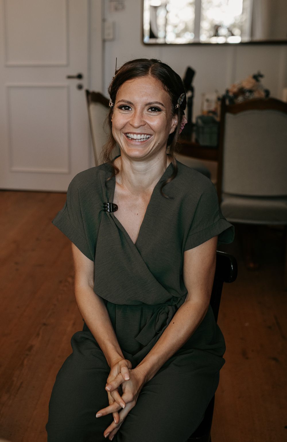 Smiling woman in green jumpsuit sitting in a bright room with wooden floors.