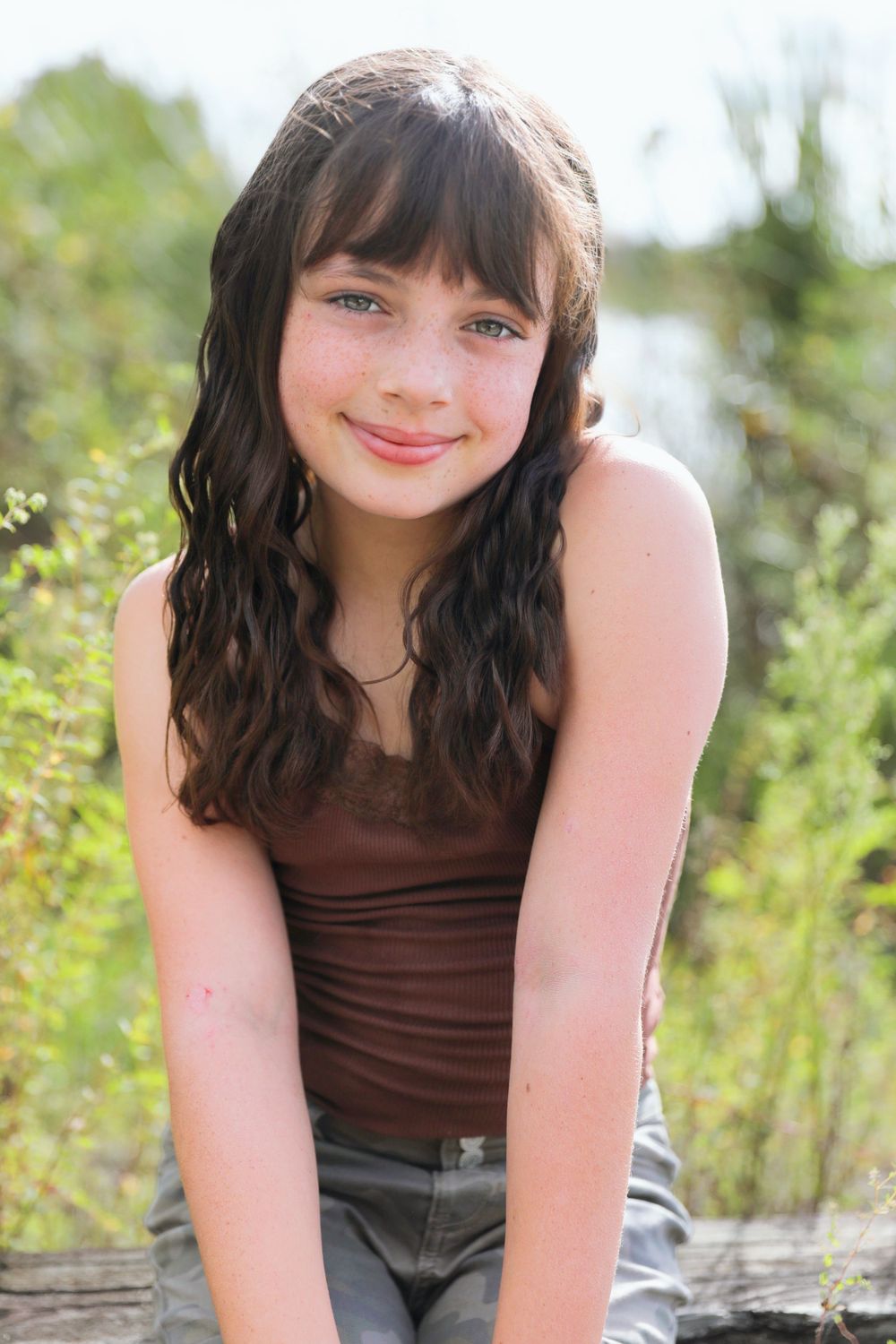 Person in a brown tank top smiles while posing outdoors on a sunny day.