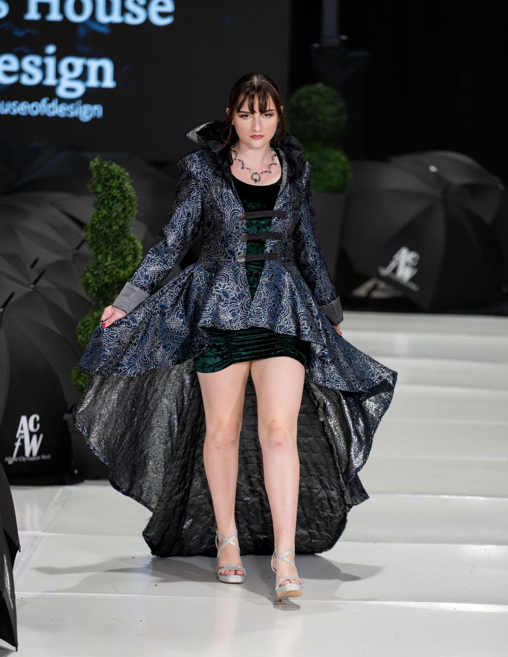 Model walks runway in dramatic navy blue metallic gown with high-low hemline at fashion show.