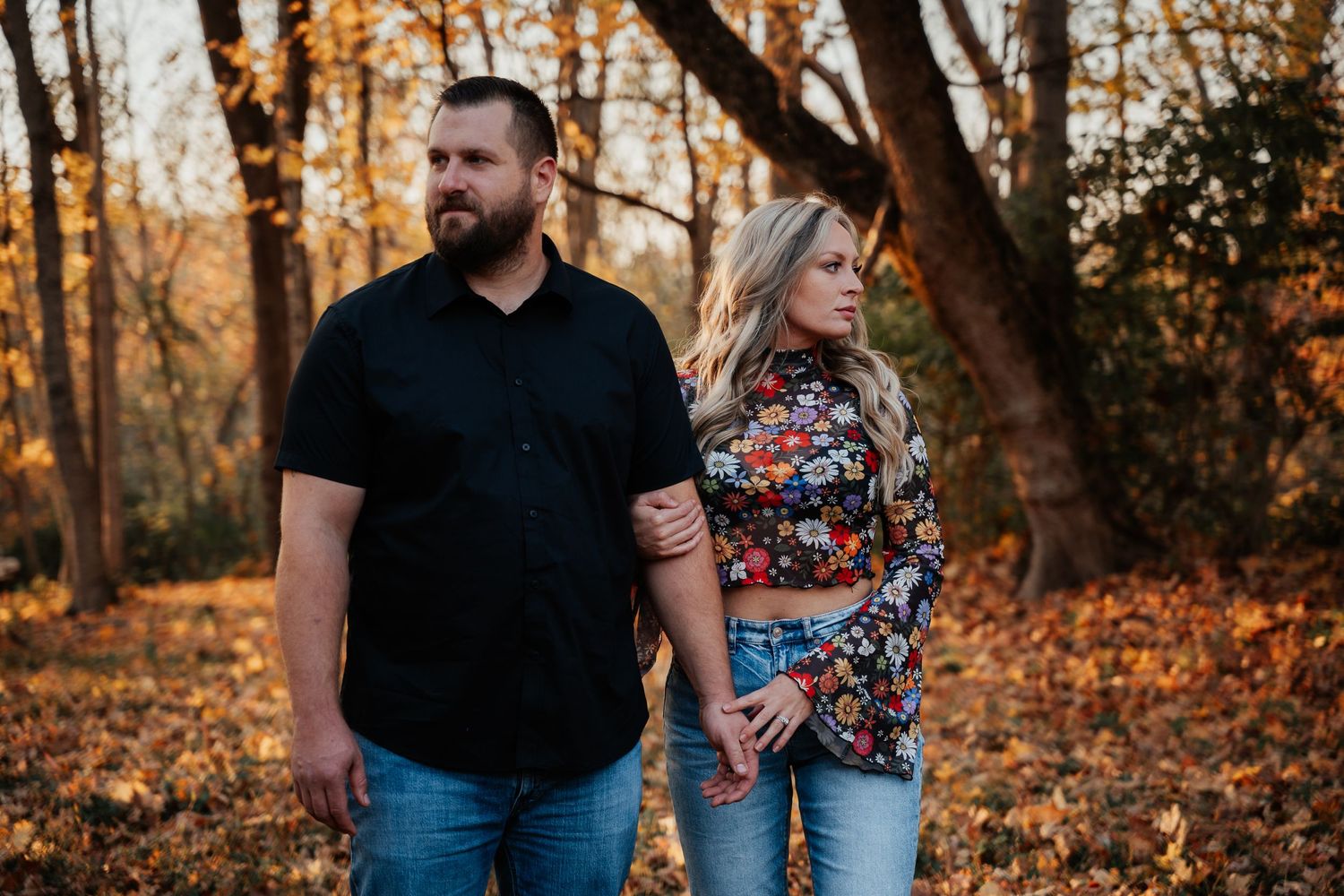 A couple poses casually against autumn trees during golden hour.