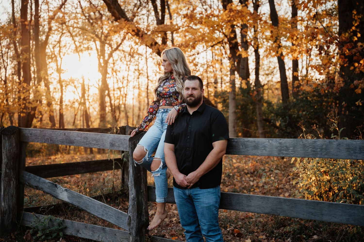 A couple poses by a wooden fence during sunset in an autumn forest setting.