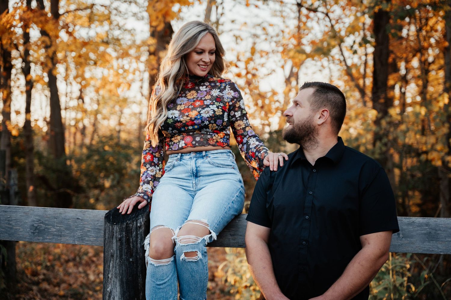 A casual couple shares a moment by a rustic wooden fence in autumn scenery.